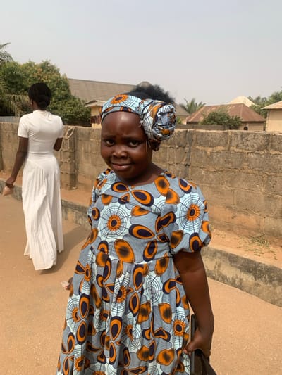 An outdoor scene in Garaku, Nigeria, features two females. In the foreground, a young female with dark skin wears a light blue dress patterned with orange and dark blue concentric circles and elongated shapes, featuring puffed sleeves and a pleated skirt. Her head is adorned with a matching fabric headwrap styled into a prominent roll on the right side, with dark, curly hair visible. She displays a slight smile and wears small hoop earrings. Her left hand is partially visible, holding a dark object. In the mid-ground, an adult female with dark skin, wearing a long, white, high-collared dress with short sleeves and a pleated section, walks away from the camera, holding a light-colored object in her right hand. Her hair is pulled back into a bun. The ground consists of unpaved, light brown earth. A concrete block wall runs along the right side of the frame. Beyond the wall, various structures are visible, including one with a light-colored, possibly conical roof, and scattered green foliage under a clear, light blue sky.