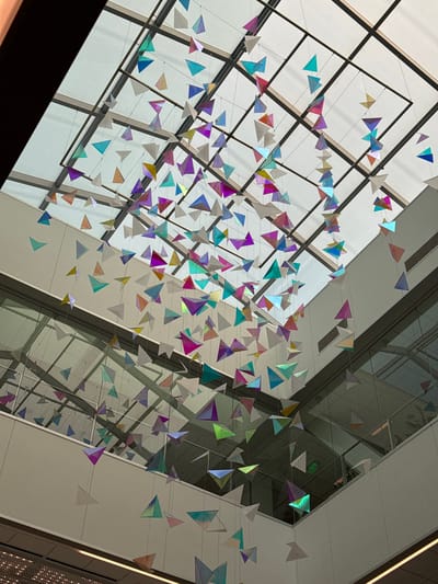 The image displays a suspended art installation featuring numerous colorful, translucent triangular prisms hanging from a grid ceiling. The prisms vary in color, including shades of blue, green, purple, pink, and yellow, and are arranged in a cascade. The ceiling appears to be made of glass panels, allowing natural light to filter through. Below the art installation, there are multi-level interior structures, including balconies with glass railings and what appear to be offices or common areas. A green "EXIT" sign is visible on one of the interior walls. The overall setting suggests an indoor public space, possibly an atrium or a lobby of a modern building. This artwork is located in the Marin County Civic Center in San Rafael, California, United States of America. There are no people clearly visible in the image.
