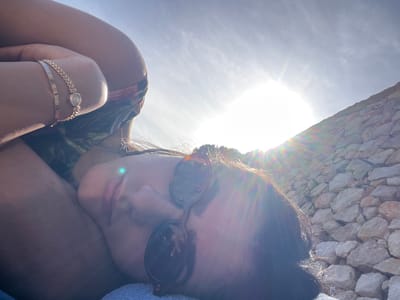 A woman wearing sunglasses and bracelets is lying on her back, looking at the camera. The sky is visible above her. She is near a stone wall, likely near the shore. Based on the location of Saint-Jean-Cap-Ferrat, France, the scene is likely outdoors on a sunny day, possibly at a beach or near a building with stone construction. The woman is wearing a patterned top, and one of the bracelets appears to be a watch.
