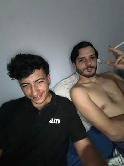 Two men pose in a horizontal selfie while lying down. One is shirtless with a beard and dark hair, appearing to hold a cigarette. The other wears a dark shirt with a white design and smiles at the camera. They rest on what appears to be a white pillow and blue bedding. The background is a plain grey wall. The picture is taken at an angle.