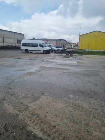 The image shows a wet, paved lot in Ovcharovo, Bulgaria, with several white vans parked in a row. A yellow industrial building is visible in the background, along with other buildings. The sky is cloudy. The ground surface appears to be a mix of concrete and asphalt with water puddles.