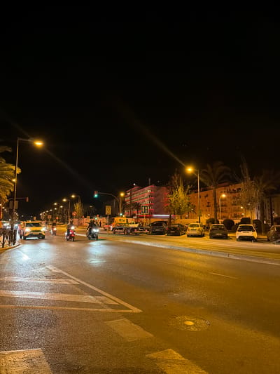 This is a nighttime, eye-level, vertical shot of a street scene in Ibiza, Spain. The image captures a road with cars and scooters, lined with palm trees and buildings illuminated by streetlights and building lights.

In the foreground, the wet asphalt road reflects the lights from street lamps and vehicles. White road markings, including a pedestrian crossing, are visible. A few scattered leaves are on the road surface.

On the left side of the road, a yellow car with license plate "EJ394 WRP" is heading towards the viewer. To its right, several scooters are traveling in the same direction, with riders wearing helmets. One scooter is red, and another is black.

Further down the road, various cars are parked along the right sidewalk. These include a yellow car and several dark-colored vehicles. The license plates of some parked cars are partially visible.

The background features multi-story buildings with balconies, their windows and facades illuminated with warm light and some red accents. A traffic light is visible, currently showing green. Palm trees and other vegetation line the street, adding to the tropical atmosphere. The sky is dark, indicating it is nighttime. A yellow tow truck is also present on the right side of the road.