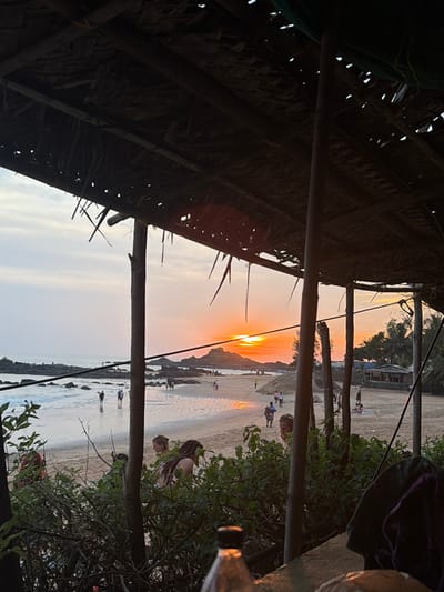 The image depicts a sunset view of a beach in Gokarna, India, observed from under a dark, thatched roof structure with prominent wooden support poles. The setting sun, an orange orb, is positioned above the horizon in the mid-ground, partially concealed by dark rock formations in the Arabian Sea. The sky displays an orange-yellow gradient near the horizon, fading to lighter tones overhead.

The sandy beach is populated by multiple individuals. Figures are seen walking along the water's edge, standing in the shallow waves, and two individuals are on horseback in the mid-left background. Distant beach shacks with thatched roofs are visible on the far right shoreline, interspersed with palm trees.

In the foreground, partially obscured by green foliage, a person with long, dark hair is depicted bending or seated. To their right, two other individuals with lighter hair are partially visible, facing towards the beach. A clear plastic bottle with a dark cap is present on a surface in the bottom-center, and dark fabric is visible on the bottom-right foreground.
