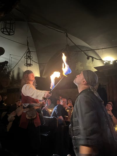The image depicts a fire performance within a dimly lit, vaulted medieval-themed restaurant in Prague, Czechia. In the foreground, two performers engage with fire. A performer on the left, with long hair and a dark bandana, dressed in a white shirt and red tunic with a leather pouch, holds two flaming torches. One torch exhibits a yellow flame, while the other displays a prominent blue and yellow flame. To their right, a second performer, wearing a dark jacket and bandana, has their head tilted back with an open mouth, directly interacting with the blue-tinged flame, suggesting a fire-eating or similar act. The restaurant interior features a dark, arched ceiling from which decorative metal cages and chains are suspended. In the background, blurred patrons are seated at tables, some holding mobile phones to capture the performance, illuminated by subtle ambient lighting.
