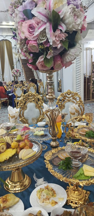 The image depicts a lavishly decorated banquet table, likely set for a celebration or wedding reception in Vabkent, Uzbekistan. A large, ornate floral arrangement dominates the foreground, featuring an abundance of pink and white roses and lilies. The table is draped in a deep blue cloth, adorned with golden trim and elaborate serving dishes.
Various food items are artfully arranged on golden platters, including a fruit platter with pineapple, bananas, apples, and grapes, as well as platters with sliced meats and what appears to be a sauce or dip. Small white plates with appetizers or side dishes are also visible. The setting includes ornate, gold-trimmed chairs with white upholstery, suggesting a formal occasion.
In the background, several people are seated at tables, engaged in conversation and enjoying the event. The lighting suggests it is likely evening. Architectural details, such as a staircase and draped curtains, hint at an indoor venue. A timestamp in the lower left corner reads "2026/06/22 20:55", indicating the date and time the photo was taken. The overall ambiance conveys a sense of festivity and elegance.