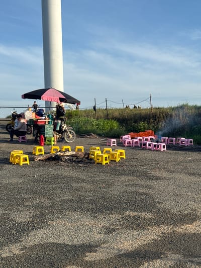 In Trường Thọ, Vietnam, an outdoor scene unfolds with a large, white cylindrical structure, possibly a wind turbine, dominating the background against a clear blue sky with scattered clouds. In the mid-ground, a mobile food stall is set up under a black and pink umbrella, equipped with various items. Two motorcycles are parked nearby, one of which is being ridden by a person. A woman is seated on a small, purple plastic stool in front of the stall, while another person stands behind it.
Scattered throughout the foreground and mid-ground are numerous small, brightly colored plastic stools, predominantly yellow and pink. Some appear arranged in seating areas, while others are dispersed on the asphalt surface. In the mid-ground, near the food stall, a small fire is burning, producing a plume of smoke. A pile of logs is visible near the fire. Beyond the stall and seating area, a wire fence runs along a grassy embankment. Several figures are seen in the distance, standing or walking near the fence line. The overall impression is a roadside gathering or market setup, likely catering to visitors or passersby.