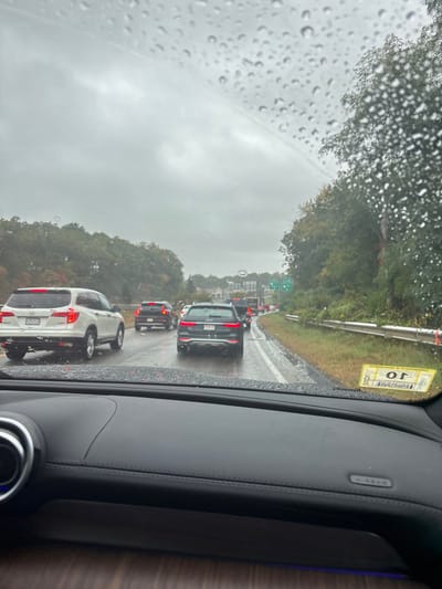 The image presents a forensic view from inside a vehicle, looking forward through a rain-splattered windshield at a traffic-congested multi-lane highway under an overcast sky. The scene is situated in Bourne, Massachusetts, United States, identified by a visible Massachusetts license plate on a vehicle and a state inspection sticker on the capturing vehicle's windshield.

The immediate foreground shows the vehicle's black leather dashboard with contrasting stitching and a dark wood-grain trim. A circular chrome-accented vent is visible on the lower left of the dashboard. An inverted yellow Massachusetts vehicle inspection sticker is adhered to the lower right corner of the windshield.

The highway is wet from rain, and several vehicles are visible in the two forward lanes. In the left lane, a white Honda Pilot SUV (Massachusetts license plate: 1XJV 72) is stationary or moving slowly, with its red taillights illuminated. To its right, in the adjacent lane, a dark gray/black Audi SUV is also visible, displaying illuminated red taillights. Further down the road, a dense line of vehicles, including larger trucks or buses, extends into the distance, indicating significant traffic congestion.

The environment alongside the highway features dense tree lines on both sides. The trees exhibit a mix of green and autumnal reddish-brown foliage. On the right side of the road, a metal guardrail separates the highway from grass and brush, beyond which more trees are present. A green overhead road sign structure is faintly visible in the far distance on the right. The overall atmosphere is dim due to the heavy cloud cover and ongoing precipitation.