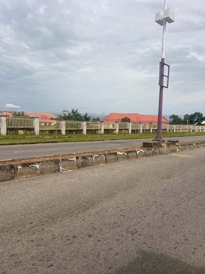 The image depicts a street scene in Kona Garu, Nigeria, under a cloudy sky. In the foreground, a paved road runs horizontally across the frame. The road has a concrete curb with visible wear and tear. Adjacent to the road is a strip of overgrown grass. Beyond the grass is a light yellow metal fence, supported by concrete posts. Behind the fence, buildings with prominent red roofs are visible. A tall purple pole with a white top and some attached equipment stands on the right side of the frame, near the road. The area appears to be a perimeter fence of a compound or facility.