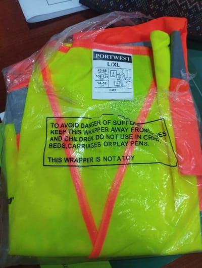 The image shows a yellow safety vest with two orange reflective stripes, folded and packaged in a clear plastic bag. It is labeled "PORTWEST L/XL," and the size specifications are listed in inches and centimeters. The vest is made of a reflective material with a bright yellow base color, and the label also includes a warning message in capital letters about the danger of suffocation and instructions to keep the wrapper away from babies and children. The location is Ashaka, Nigeria, at coordinates 10.929° N, 11.476° E.