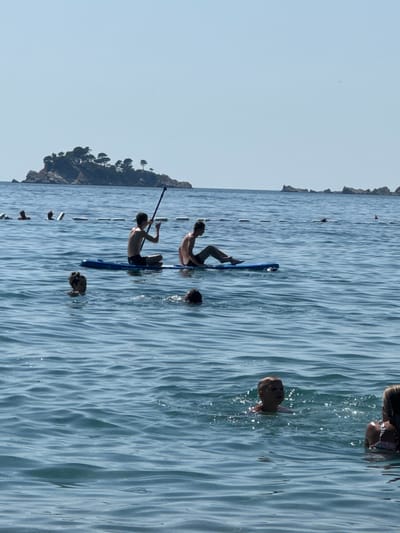 In the waters off the coast of Đurovići, Montenegro, two young males are seen on a paddleboard. The paddleboarder on the left is seated and holds a paddle, while the one on the right is lying down. Several other individuals are in the water, swimming or wading. In the background, a small, vegetated island rises from the sea, and the sky is clear and blue. A marker buoy with a red top is visible further in the distance. The water is calm with gentle ripples, reflecting the bright daylight.
