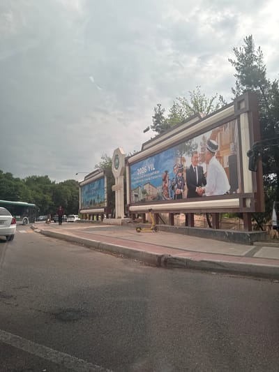 This photograph depicts a street scene in Salar, Uzbekistan. The image captures a roadside view, where a series of billboards are lined up along the sidewalk. The billboards display images of people, local architecture, and text. The text in one billboard reads, "2026 YIL MAHALLANI RIVOJLANTIRISH VA JAMIYATNI YUKSALTIRISH YILI". Cars and other vehicles, including a bus and a white car, are visible on the asphalt road adjacent to the sidewalk. Trees line the backdrop, adding to the urban environment. The sky is overcast, with clouds filling the majority of the frame. The perspective of the photo is somewhat tilted to the side.