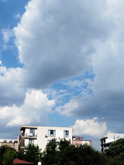 The image depicts a cloudy sky over a residential area in Bucharest, Romania. The sky is filled with a mix of large, fluffy white cumulus clouds and darker, grayer storm clouds, suggesting a dynamic weather pattern. Patches of bright blue sky are visible between the clouds.
In the foreground, the tops of several modern apartment buildings are visible, peeking out from behind lush green trees. The buildings have flat roofs and feature balconies with dark railings. One building has a banner hanging from a balcony that appears to have text on it. The time of day is likely late morning or afternoon, given the bright but diffused sunlight. The overall mood is somewhat dramatic due to the heavy cloud cover, but the presence of buildings and trees indicates a populated urban environment.