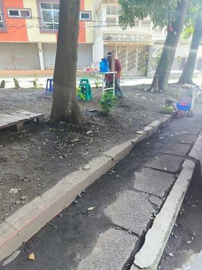 The scene takes place in an urban area in Kecamatan Dayeuhkolot, Indonesia, with coordinates -6.965 latitude and 107.59 longitude. A single person, wearing a red shirt and black pants, sits at a table with a water cooler and a few small objects on it. There is a pallet on the ground to the left of the table, with a tree trunk and roots in the foreground. Two blue plastic stools are nearby, and a small plant is growing in the ground. The ground is covered with a layer of dirt and small pebbles. A concrete curb borders a paved sidewalk. The environment is dry and dusty. There are several buildings in the background, obscured by tree foliage. One of the trees is large and its trunk is visible on the left of the image. The sidewalk is cracked and uneven. A thin cable runs overhead. The overall scene is one of casualness and routine activity.