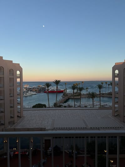 This image depicts a hotel balcony view overlooking a marina and the Red Sea in Hurghada, Egypt. The scene is set during dusk, with a clear blue sky transitioning to a warm, soft orange and yellow gradient near the horizon, indicating sunset or sunrise. The moon is visible as a small, bright orb in the upper left quadrant of the sky.

On the balcony, white metal railings create a barrier in the foreground. The balcony floor is paved with a light-colored mosaic tile pattern. Beyond the balcony, a body of water, likely the Red Sea, is filled with numerous boats and yachts docked in a marina. The water appears calm and reflects the soft light of the sky.

Several palm trees are strategically placed along the waterfront, their fronds visible against the backdrop of the sea and sky. A prominent red and white boat, likely a ferry or larger yacht, is docked near the shore, adding a splash of color to the scene.

In the background, the distinct architecture of resort buildings is visible, characterized by their pale facades and multi-story structures with balconies and windows. The image captures a tranquil and inviting atmosphere, typical of a resort destination. There is no visible text in the image.