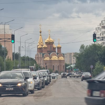 The image captures a street scene in Aktobe, Kazakhstan, dominated by traffic and a prominent Orthodox church with golden domes. The perspective is from a vehicle, looking forward along a paved road with visible wear and tear. A line of cars is stationary or moving slowly in the foreground and middle ground. On the left side of the road, a traffic light displays a green signal. Beyond the traffic, a large, ornate church with multiple gold-plated domes and crosses stands as the focal point. The architecture suggests a significant religious structure. In the background, apartment buildings and trees line the sides of the road, indicating an urban or suburban environment. The sky is overcast with heavy gray clouds. A few streetlights are visible along the road. The overall atmosphere is one of a typical city day, with moderate traffic and a striking architectural landmark.