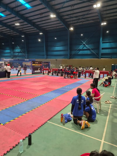 The image depicts an indoor sports arena, likely a gymnasium, in Phagwara, India, filled with participants and spectators for a sports event, possibly a kabaddi match given the branding on a banner. The floor is covered with interlocking foam mats in red and blue, separated by yellow and black striped tape. Several individuals, predominantly young women in athletic attire, are visible on and around the playing area, some actively participating, others resting or observing. Banners displaying event information, including "Kendriya Vidyalaya Sangathan 60th National Sports Meet 2023," are displayed on the back wall. Spectators are seated on chairs along the sidelines, and some individuals are standing, observing the proceedings. The arena is illuminated by overhead lights, and the ceiling structure is visible. The overall atmosphere suggests a competitive sporting event in progress.