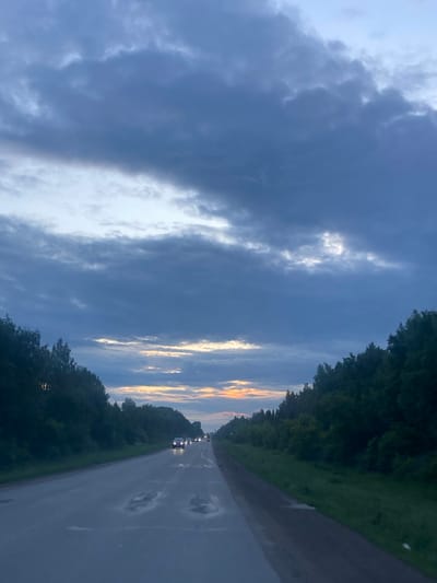 The image depicts a road scene at dusk, with a focus on the sky and the surrounding environment. The sky is filled with dramatic, dark clouds, interspersed with streaks of pale light, suggesting the last moments of daylight. A sliver of golden and orange light is visible on the horizon, hinting at a sunset.
The road itself appears to be wet, with several puddles reflecting the dim light, indicating recent rain. Trees line both sides of the road, forming a dense, dark border that frames the scene. In the distance, several cars are visible on the road, their headlights on, suggesting ongoing traffic.
The overall mood of the image is somewhat somber and serene, with the twilight setting and the wet road contributing to a feeling of quiet transition. The location context provided is Nurkeevo, Russia. There is no visible text in the image.