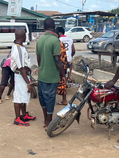 The image captures a street scene in Ijegun, Nigeria. A red motorcycle is parked on the side of the road. Several people are standing nearby. One person is wearing a green shirt and dark shorts. Another person is wearing a white and black shirt, white shorts, and red and black sneakers. In the background, there are buildings, cars, and stacks of blue crates.