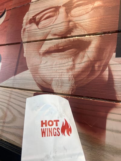 A white paper bag with "HOT WINGS" printed in red and a flame graphic is held in the foreground, obscuring part of a large, close-up image of Colonel Sanders' face printed on a wooden background. The image of Colonel Sanders is faded and appears to be part of an advertisement or mural. The wooden background has horizontal planking. The scene is outdoors and appears to be a bright day, with shadows present. The location is identified as a KFC restaurant in Manchester, United Kingdom.