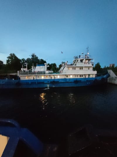 A blue and white vessel, possibly a tugboat, is docked on a dark body of water. The vessel has multiple windows illuminated from within. The sky above is a deep twilight blue. Trees line the bank behind the vessel. The scene appears to be in the city of Sheksna, Russia, on a canal or river.