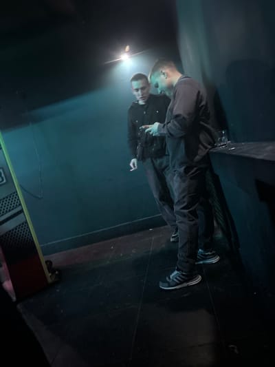Two young men, appearing to be in their late teens or early twenties, stand in a dimly lit, indoor space with teal-colored walls. The man on the right, facing the viewer's left, is looking down at his phone, which he holds in his right hand. He is wearing a dark-colored, long-sleeved shirt and dark pants, with athletic shoes. The man on the left, facing the viewer's right, is looking at the phone as well and appears to be holding a cigarette in his left hand. He is wearing a dark shirt and dark pants. A dark counter or table is visible to the right of the two men, with a glass and possibly other items on it. A piece of equipment with a yellow and red frame is partially visible on the left side of the image. The lighting suggests a club or concert venue setting. The location in the city of Kirov, Russia, is not discernible from the image.