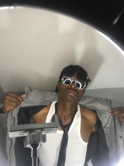 The image depicts a young male individual, located in Kirana, Nigeria, posing indoors for a portrait. The subject, of dark complexion, faces directly forward, wearing dark sunglasses with square metallic frames. He has short, dark, textured hair. He is dressed in a white tank top, over which a dark-colored tie is loosely draped around his neck, hanging centrally. Both hands are visible, holding open a light gray jacket on either side of his torso, revealing the inner lining and the tank top. In the immediate foreground, a black and gray smartphone holder, attached to a tripod or stand, is positioned centrally below the subject's chest. The background consists of a plain, light-colored wall. Artificial lighting, originating from a ring light, is evident around the upper perimeter of the frame, casting a bright, even illumination on the subject and background.