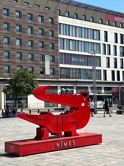 This is a vertical, outdoor, daytime photograph taken in Nîmes, France. The scene features a large, abstract red sculpture in the foreground. The sculpture appears to be made of metal and has a jagged, claw-like shape. To the left of the sculpture is a tall, modern building with a facade of windows and vertical wooden slats. Another, taller building with a concrete facade and numerous windows is visible behind the red sculpture. A tree with green foliage stands between the buildings.

Several people are present in the scene, mostly in the background, appearing to walk or sit on a paved area in front of the buildings. The overall atmosphere is sunny and bright, suggesting a pleasant day.

Visible text includes "NÎMES" in large white letters on the side of the red sculpture. On the buildings, there are signs and window markings that read "Midi Libre" and "MAISON de MA," "ibis STYLES," and "HOTEL." The presence of these signs, along with the architectural style, suggests an urban setting, possibly a commercial or residential area. The red sculpture is the dominant object, and its abstract nature draws the viewer's eye, contrasting with the more geometric forms of the buildings.