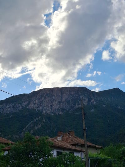 The image, oriented vertically but depicting a landscape scene, captures a dramatic mountainous environment under a partly cloudy sky. The left side of the frame is dominated by the sky, featuring large, white and grey clouds interspersed with clear patches of blue, indicative of a daytime scene with mixed weather.
The right side of the image showcases a prominent, steep mountain. Its lower slopes are densely covered with dark green trees and foliage, while higher sections reveal rugged, rocky terrain. Towards the bottom right corner, a portion of a building with a traditional terracotta tiled roof is visible, partially hidden by verdant trees. A tall, dark wooden utility pole stands out in the mid-right, with multiple power lines extending from it and stretching across the scene. Other lines are also seen traversing the foreground, suggesting an electrical or communication infrastructure in the area.
There are no people or explicit activities taking place in the image. The scene presents a tranquil, natural setting with subtle hints of human habitation and infrastructure, characteristic of a rural or semi-rural area. Given the context, this landscape is located in Lyutibrod, Bulgaria. No visible text is present in the image.