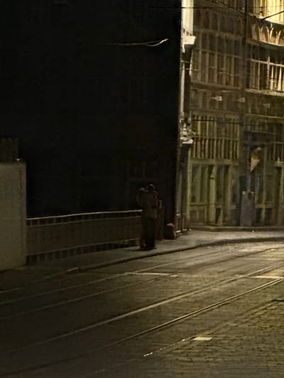 The image depicts a nighttime scene on a cobblestone street with tram tracks in Ghent, Belgium. On the left side of the image, a dark building with a light-colored vertical element is visible. A low railing runs along the edge of this area. In the mid-ground, two figures stand near a dark object on the ground. To the right, a multi-story building with numerous windows and decorative architectural features is illuminated. The street surface is paved with cobblestones and features visible tram tracks. The overall lighting is dim, with artificial light emanating from the building on the right and a hint of light in the sky.