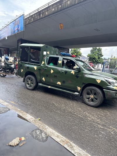 This image depicts a scene in Ikoyi, Nigeria, likely during the day under overcast skies, as indicated by the wet road and reflections. The primary focus is a green, camouflage-patterned pickup truck, likely a security vehicle, with a red and blue light bar mounted on its roof. The truck has a reinforced rear section, possibly for carrying personnel or equipment. The side of the truck features a prominent "GUINNESS" logo, suggesting a sponsorship or advertising presence.
In the foreground, a motorcycle with a rider wearing a blue helmet is partially visible, hinting at the common mode of transport in the area. The road surface appears wet and somewhat dirty, with visible tire tracks and scattered debris. Overhead, a large concrete overpass or bridge dominates the upper portion of the frame, casting a shadow. Below the overpass, a billboard advertising "FOR MORE THEN YOU ARE!" with images of people is mounted. Another sign for "BONNY CANTONMENT" and "NAVAL DOCTRINE COMMAND" is visible on a building in the background, suggesting a military or naval presence in the vicinity. The overall atmosphere appears to be a busy urban environment, possibly during or after a rainfall.