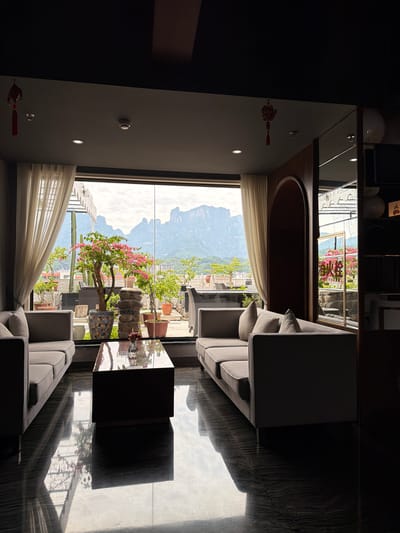 A bright, daylight interior view reveals a living space overlooking a cityscape. The room contains two gray sofas and a dark-colored coffee table, positioned on a shiny, dark-toned floor reflecting sunlight. A large window provides the primary source of natural light and offers an expansive view of mountains, potentially near the city of Zhangjiajie in China, as well as nearby buildings with flora. There are white sheer curtains on the edges of the window. On the interior walls, decorative Chinese knots can be observed. Above the window, a shelf is visible. The scene captures a balance of indoor comfort and an engagement with the outdoor landscape.