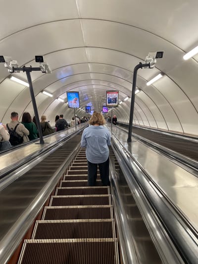 The image shows a wide, downward-sloping escalator in a subway station, likely in Kipen, Russia, given the context. The escalator is surrounded by a white, arched tunnel with rows of fluorescent lights and surveillance cameras mounted on black poles. Several people are visible on the escalator, mostly facing away from the camera and ascending. A woman in a light blue striped shirt and dark pants stands prominently on the escalator in the foreground, facing away from the camera and looking down the steps. To her left, further down the escalator, other individuals are seen in profile, some carrying backpacks. Posters are displayed on the tunnel walls at intervals. The escalator steps are dark brown with lighter yellow risers. The overall impression is one of a busy public transportation hub.