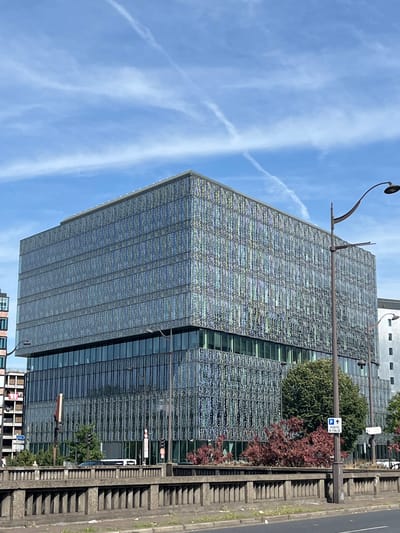 The image shows a modern, multi-story building with a distinctive facade featuring a repeating geometric pattern of metallic vertical elements. The building has a glass lower level and appears to be in an urban setting. In the foreground, there is a concrete barrier with vertical supports, suggesting it is a bridge or elevated roadway. Streetlights are visible, one with a curved arm. Trees and some red foliage are present behind the barrier, in front of the building. A blue and white parking sign with an arrow pointing upwards and the text "PORTE D'ITALIE" is affixed to a lamppost. The sky is blue with scattered white clouds and contrails from an aircraft. To the left of the main building, another building with balconies is partially visible. The location is Le Kremlin-Bicêtre, France.