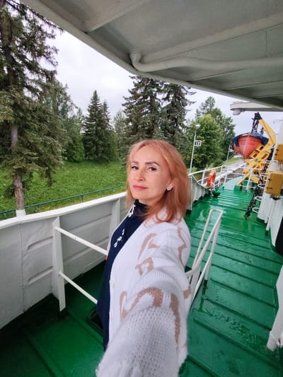 A medium shot captures a woman with reddish-blonde hair, wearing a white knitted cardigan over a dark top, standing on the deck of a green-painted boat. She is facing the camera and appears to be taking a selfie. The background shows lush green trees and foliage, suggesting an outdoor, possibly rural or semi-rural location. Part of the boat's structure, including railings and equipment like a life raft and a crane, is visible. The deck appears wet, indicating recent rain or spray. The image is taken from a slightly elevated perspective. The presence of a life raft and crane suggests the boat is a working vessel, possibly used for transport or other maritime activities. The city of Белоусово, Russia, is mentioned in the prompt but there are no direct visual cues in the image to confirm this specific location.