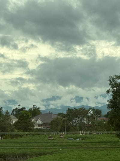 The image depicts a rural landscape in Hai Lang Commune, Vietnam. The foreground is dominated by a vibrant green field, possibly a rice paddy, crisscrossed by dark horizontal lines, suggesting irrigation channels or power lines. In the mid-ground, several houses with tiled roofs are nestled amongst lush green trees, partially obscured by foliage. These homes appear to be of traditional Vietnamese architecture. Beyond the houses, a range of mountains looms in the distance, their peaks shrouded in mist and clouds, creating a hazy, atmospheric effect. The sky is overcast with heavy, gray clouds, indicating a potential for rain or a continuation of dull weather. The overall scene is one of tranquility and natural beauty, characteristic of rural Vietnam.