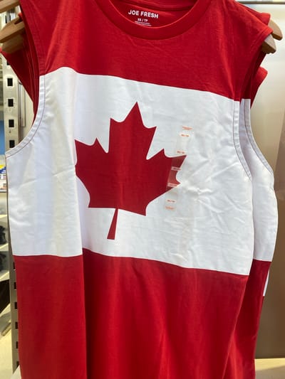 The image shows a rack of sleeveless red and white t-shirts featuring the Canadian flag design, hanging on wooden hangers in a retail store. The t-shirts are in the foreground, with a rack of clothing visible in the background to the left. The lighting appears to be artificial, consistent with an indoor retail environment. No people are visible in the image, and no specific location within Whitehorse, Canada, can be determined from the visual information provided. The t-shirts have size tags attached.