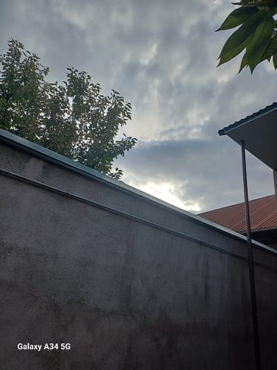 This is a low-angle, vertically oriented outdoor photograph taken in Angren, Uzbekistan. The scene captures a cloudy sky with patches of sunlight breaking through, suggesting it might be late afternoon or early morning.
The dominant subjects are the sky, clouds, and portions of a building's exterior. On the left side, the foreground features large green leaves of a tree, with some appearing slightly wilted. Beneath the leaves, a portion of a building's angled roof with a greenish trim is visible. The main body of the building, to the right, is a textured, grey wall. A thin, dark pipe runs vertically along this wall.
Further back, another section of a building with a reddish, corrugated roof is visible, separated by a horizontal dark pipe. The overall impression is one of an overcast sky with intermittent sunlight, illuminating the scene. There are no people or animals present. The texture of the wall and the natural elements of the tree leaves and sky contribute to the visual composition.
The visible text on the bottom right of the image reads "Galaxy A34 5G", which is a device watermark.
