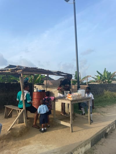 An outdoor market stall is depicted on a dirt path in Mosafejaw, Nigeria, under a light blue sky with scattered clouds. The stall consists of a rough wooden frame supporting a corrugated metal sheet roof. On the left, a woman in a teal shirt with a white cloth draped over her shoulder sits on a wooden bench. Adjacent to her is a red metal barrel topped with a cooking pot. A second woman, identifiable as a vendor, stands behind a wooden table displaying packaged bread, a clear plastic container, and a dark, unidentified pile. An infant is secured to her back. Three additional children are present: a boy in a light blue school uniform and dark shorts stands near the table, facing it; a girl in a pink outfit with a backpack stands slightly behind him; and a second boy in a white school shirt and plaid shorts stands to the right of the table, holding a white plastic pitcher. A tall, dark grey solar-powered streetlight pole is centrally located behind the stall. The background features a low concrete wall, a corrugated metal fence, and dense green foliage, including banana trees.