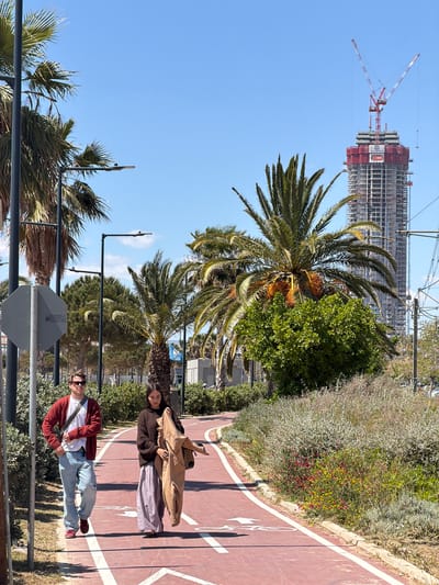 On a sunny day in Glyfada, Greece, a man and woman are visible on a red paved path bordered by greenery. The man, facing the camera, wears a red cardigan over a white shirt, light blue jeans, and red shoes. He has sunglasses on. The woman is lying down. She is wearing a dark jacket and light-colored skirt. She has a light brown jacket on top of her and a brown bag. Behind them are palm trees. In the distance, a tall building under construction is visible. Cranes are active around the building. A signpost is visible in the foreground.