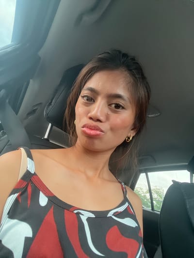 A young woman with brown hair and brown eyes is taking a selfie inside a vehicle. She is wearing a black, red, and white patterned tank top and gold hoop earrings. She is pouting with her lips. The background shows the interior of a car, including a seatbelt, headrest, and a window with trees visible outside. The image appears to have been taken during the day. The location is identified as Carmen, Philippines.