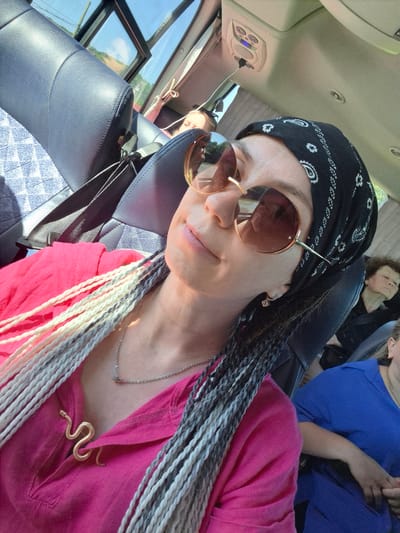 In the city of Krasnyy Klyuch, Russia, a woman with a black bandana and sunglasses takes a selfie in a bus. She is wearing a bright pink shirt with a snake-shaped brooch and has blonde and grey braided hair. The bus is traveling during the day, with sunlight streaming through the windows, illuminating the interior and casting shadows. Other passengers are visible in the background, some appearing to be sleeping. The seats are upholstered in dark blue leather, and a seatbelt is visible. The environment outside the bus suggests a rural or suburban landscape with trees and vegetation.