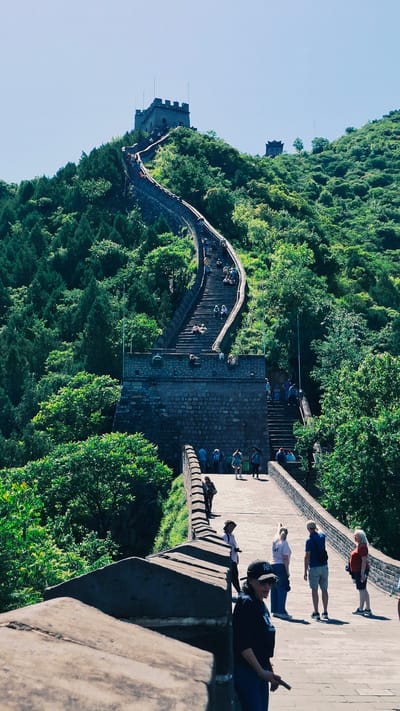 The image shows a section of the Great Wall of China in Changping District. The wall winds along the ridgeline of a heavily forested mountain. Several watchtowers are visible. Groups of tourists are walking on the wall. On a paved section of the wall, several people are standing and sitting. A clear blue sky is overhead.