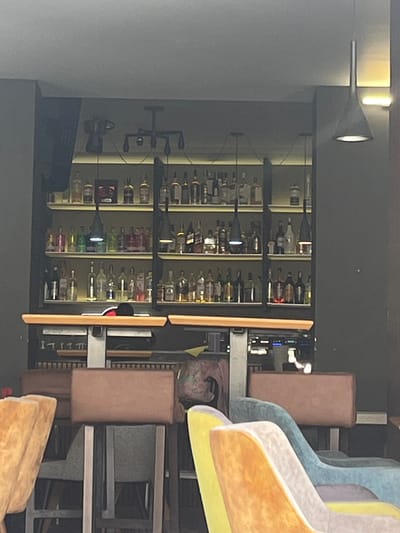 The image captures an interior scene of a bar or restaurant located in Dupnitsa, Bulgaria. The central feature is a well-stocked bar area with multiple shelves displaying a wide variety of bottles, likely alcoholic beverages. These shelves are intricately lit from behind and possibly from within, creating a glowing effect on the bottles. Several modern pendant lights hang in front of the shelves, contributing to the illumination of the bar.
To the left of the bar, a light grey wall is visible with a sleek, contemporary wall-mounted spotlight extending from it, casting light towards the scene. On the right side of the frame, a seating area is present, featuring several chairs and bar stools. The chairs are upholstered in various pastel colors, including light blue, light grey, yellow, and orange, indicating a modern design. Some bar stools with brown upholstered seats are also visible, arranged around a counter or table structure.
No people are present, and no specific activity is taking place, suggesting the establishment might be closed, about to open, or captured during a quiet moment. The lighting is artificial, giving the impression of evening or night-time. The overall ambiance appears sophisticated and welcoming. No discernible text is visible on any of the bottles or furniture.