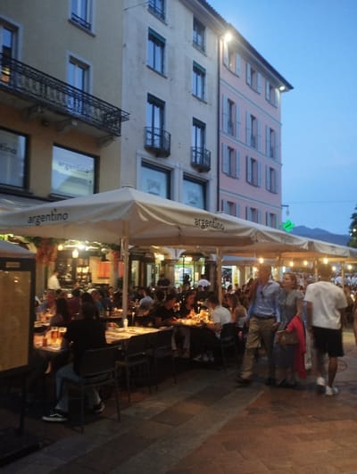 The image depicts an outdoor dining scene in Lugano, Switzerland. Several people are seated at tables under large white umbrellas, enjoying a meal. The umbrellas are branded with the word "argentino". In the background, there are buildings with windows and balconies. The sky is a dusky blue, suggesting it is either early evening or late afternoon. The lighting appears to be artificial, with string lights illuminating the dining area. A few individuals are walking through the scene, while others are seated, engaged in conversation or eating. The ground is paved with cobblestones.