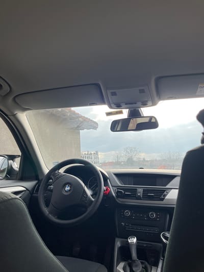The image depicts the interior of a BMW vehicle, specifically viewed from the rear passenger seat looking forward. The primary subjects are the car's cockpit and the exterior environment visible through the windshield.

**Vehicle Interior:**
The steering wheel, adorned with the BMW logo and integrated multifunction buttons, is prominent on the left, covered in black textured material. To its right, the dashboard is black, featuring a central air vent system, climate controls, and a radio unit. A manual gear stick with a silver top is located in the center console. A dark fabric seat, likely the driver's, is partially visible on the far left. The front passenger seat headrest is visible on the far right. Above, the gray headliner includes sun visors and an overhead console. A rear-view mirror is centrally positioned, reflecting a light-colored interior element. A yellow sticker with "BG 41" is affixed near the rearview mirror mount. A small red object, possibly an air freshener or accessory, is attached near the steering column.

**Exterior Environment (Sredkovets, Bulgaria):**
Through the windshield, a partial view of a building with a weathered, possibly terracotta-tiled roof is visible on the left, indicating proximity to a structure. The building wall appears light-colored. Further into the distance, a large white structure with an ornate, possibly skeletal or latticework design is discernible on the left, suggesting a modern public building or stadium. On the right, bare deciduous trees are visible against a cloudy sky, where sunlight breaks through, creating a glare on the windshield. The overall lighting suggests an overcast day with some direct sun. The scene is located in Sredkovets, Bulgaria. No people or active interactions are visible in the frame.
