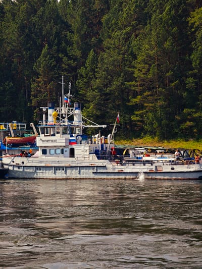 In the city of Porog, Russia, on a body of water, a white and gray boat with multiple decks and a superstructure is docked. The boat has the Cyrillic text "РОСМОРРЕЧФЛОТ" on its side. A Russian flag is visible on a mast. The boat is situated against a backdrop of a dense, green pine forest that rises from the shore. The water in the foreground shows ripples and small waves. Several people are visible on the deck of the boat, some appearing to be working. A small orange life raft is attached to the side of the boat.