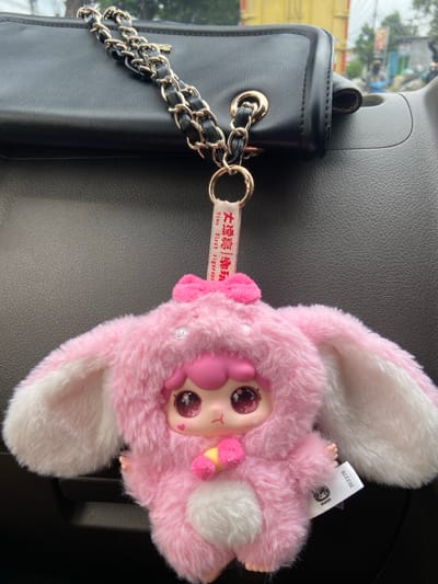 A black purse with a gold chain rests on a gray surface. Attached to the purse is a pink, fuzzy keychain featuring a character with large eyes and rabbit ears. The keychain has a tag with text in what appears to be Chinese and English. The plush keychain appears to have a fluffy tail and a small, pink and yellow accessory attached to it.