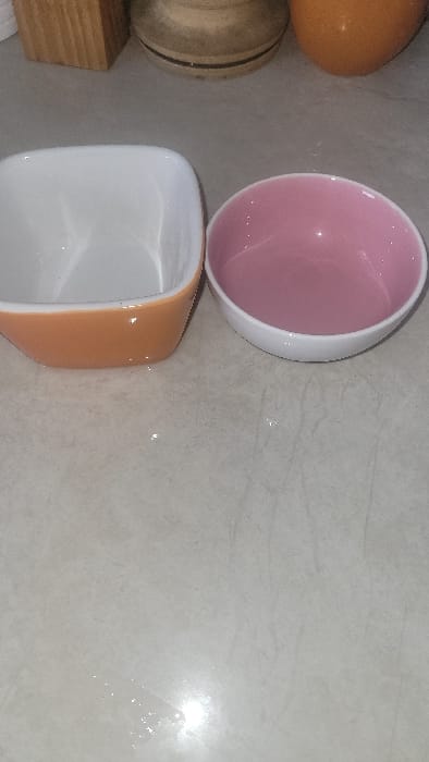 This image depicts an indoor scene in the city of Nanyuki, Kenya. In the foreground, two empty ceramic bowls are positioned on a light-colored, reflective countertop or tiled surface. The bowl on the left is square-shaped with a white interior and an orange-brown exterior. The bowl on the right is round with a pink interior and a white exterior. In the blurred upper background, portions of several objects are visible: a light brown wooden block on the left, a beige, striped, round vessel, and an orange-brown ceramic object on the right. No individuals or actions are present in the frame.