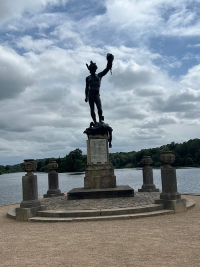 This is a vertical-format, outdoor, eye-level shot featuring a dark bronze statue of Perseus holding Medusa's head. The statue is positioned in front of a body of water and a line of trees. The sky above is filled with dramatic white and gray clouds. The statue stands on a pedestal which has a plaque with text. The plaque reads: "PERSEUS AND MEDUSA. ORIGINAL STATUE SCULPTED BY RIVINITO CELLINI IN FLENCE FOR THE GRAND DUKT OF TUSCANY MOL RIPUKA MARE GEDIE OF DUKE ONS THERLAND WITH THE FLEMISSION FRIEND THE THEN CLAND DUKE OF TUSCANY WAS ORIGINAL ALLY FRICCIO MERT ON THE ONSTRUCTION OF TRENTHAM MDCCCXL HERE RESTORED AND REPLACED HU BY KUZABETH CORINTESSO SUTHERLAND". The statue is part of a garden setting, with stone pillars and paving stones visible in the foreground. The overall impression is one of historical significance and grandeur, set against a somewhat overcast sky. The time of day appears to be daytime, and the weather is cloudy. The setting suggests a park or garden with a prominent water feature.
