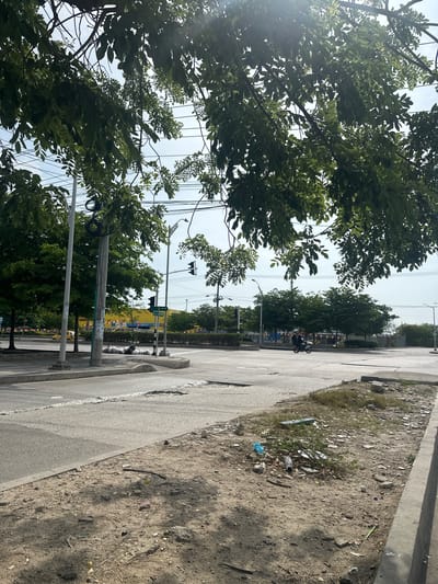 The image shows a street intersection in Perímetro Urbano Barranquilla, Colombia. A motorcycle with two occupants is crossing the intersection. Trees line the street, and electrical wires are visible overhead. The road surface is asphalt with visible wear and tear. On the right side of the road, there is a strip of unpaved ground with debris and scattered trash. The sky is overcast.